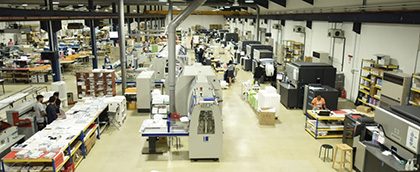 zensmart is showing A spacious, well-lit factory floor filled with various types of machinery, workstations, and shelves stocked with materials. Several workers are operating machines and organizing handmade creations, contributing to a busy atmosphere reminiscent of vintage craftsmanship and manufacturing. with print workflow automation