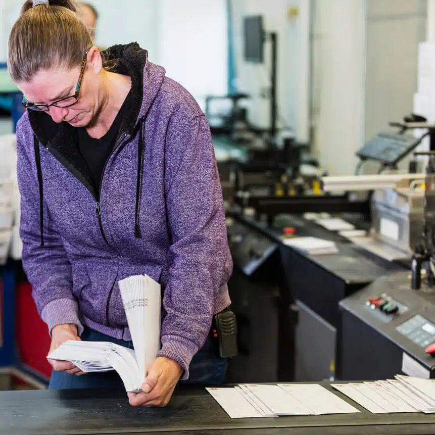 zensmart is showing A person wearing glasses and a purple hoodie reviews a stack of papers in a room with industrial printing equipment. The setting appears to be a print shop, with various printing machines and stacks of paper visible around them, likely preparing materials for direct mail campaigns. with print workflow automation