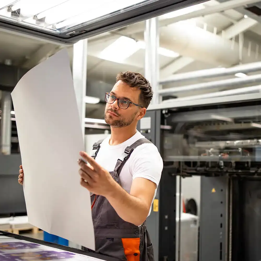zensmart is showing A man wearing glasses and overalls examines a large sheet of paper in a modern industrial setting with machinery in the background. The man is focused on the paper, possibly checking its quality or details for signage or display purposes. with print workflow automation