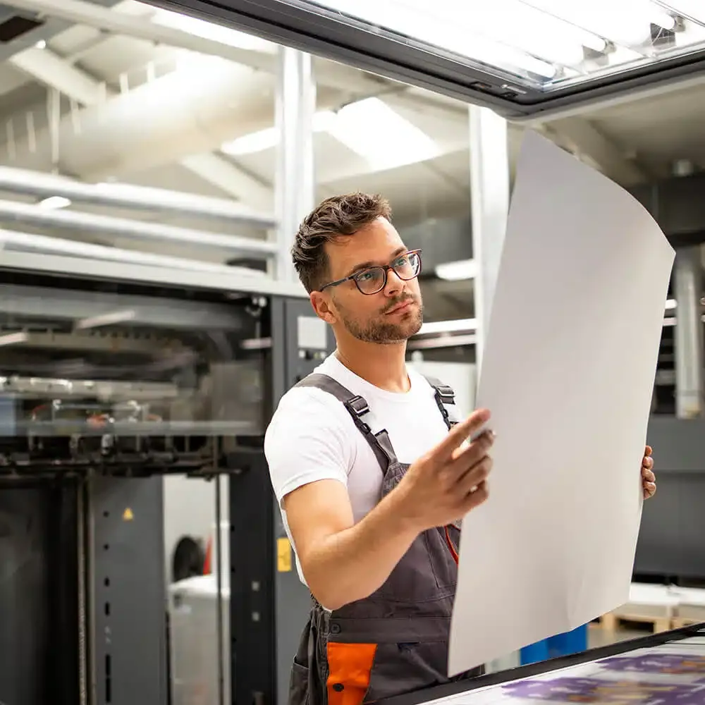 zensmart is showing A man in glasses and work overalls examines a large sheet of paper in a modern printing facility. He appears to be inspecting the quality of the print, which could be used for signage or banners. The industrial background includes printing machinery and overhead lighting. with print workflow automation