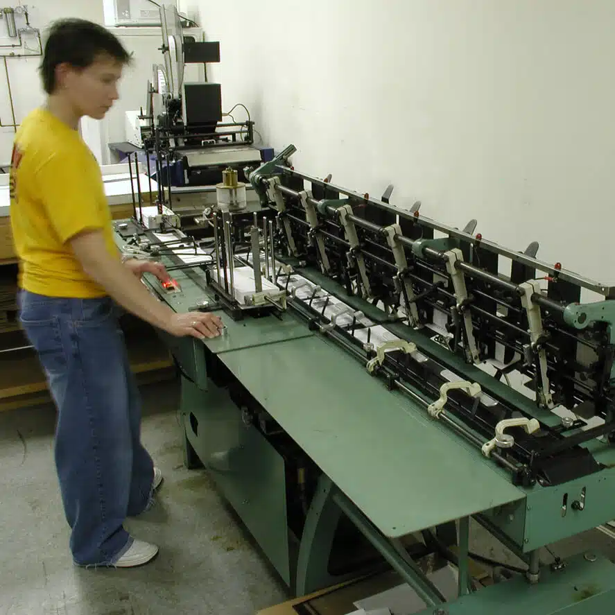 zensmart is showing A person in a yellow shirt and blue jeans operates a green industrial bookbinding machine in a workshop setting. The machine, integral for Direct Mail projects, is equipped with multiple clamps and levers. The workspace appears organized with additional equipment visible in the background. with print workflow automation