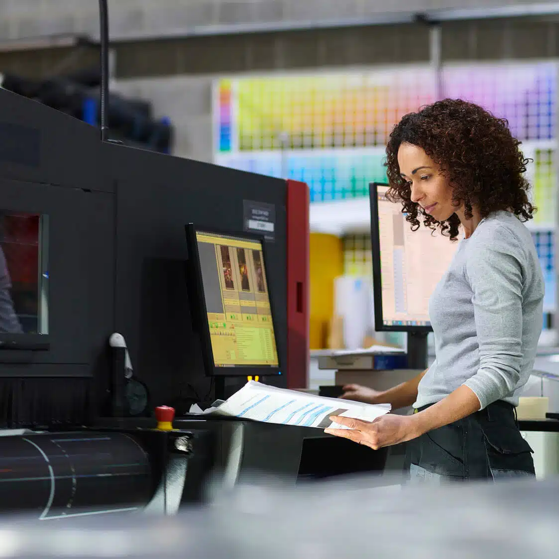 zensmart is showing A woman with curly hair stands in front of a large industrial printer, reviewing a printed document. She is wearing a light gray long-sleeve shirt. The background shows colorful photo prints and computer monitors displaying various graphics. with print workflow automation