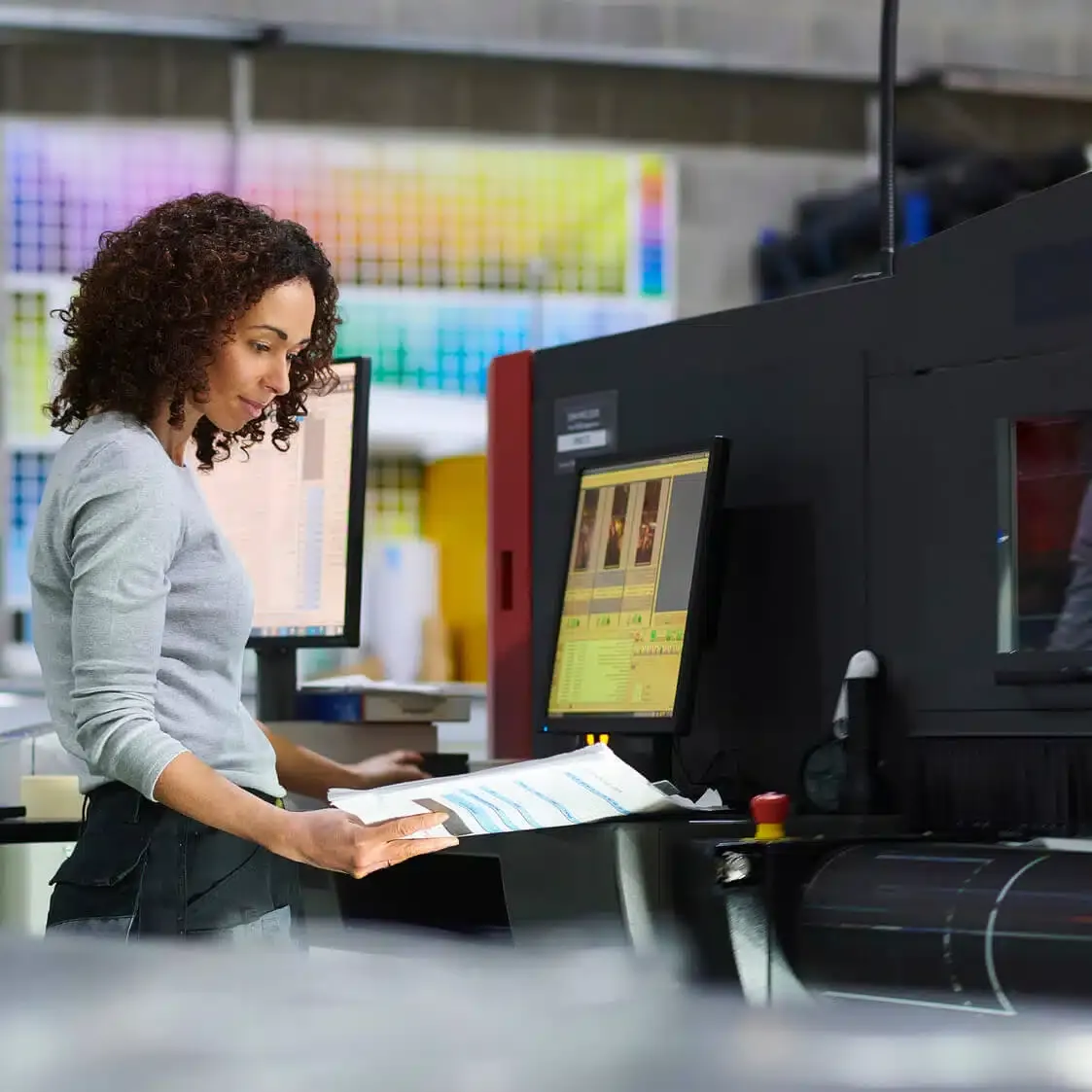 zensmart is showing A person with curly hair wearing a gray shirt stands in front of a large printer in a print shop, holding a paper and looking at a computer screen. Colorful photo prints are displayed on the wall behind them. with print workflow automation