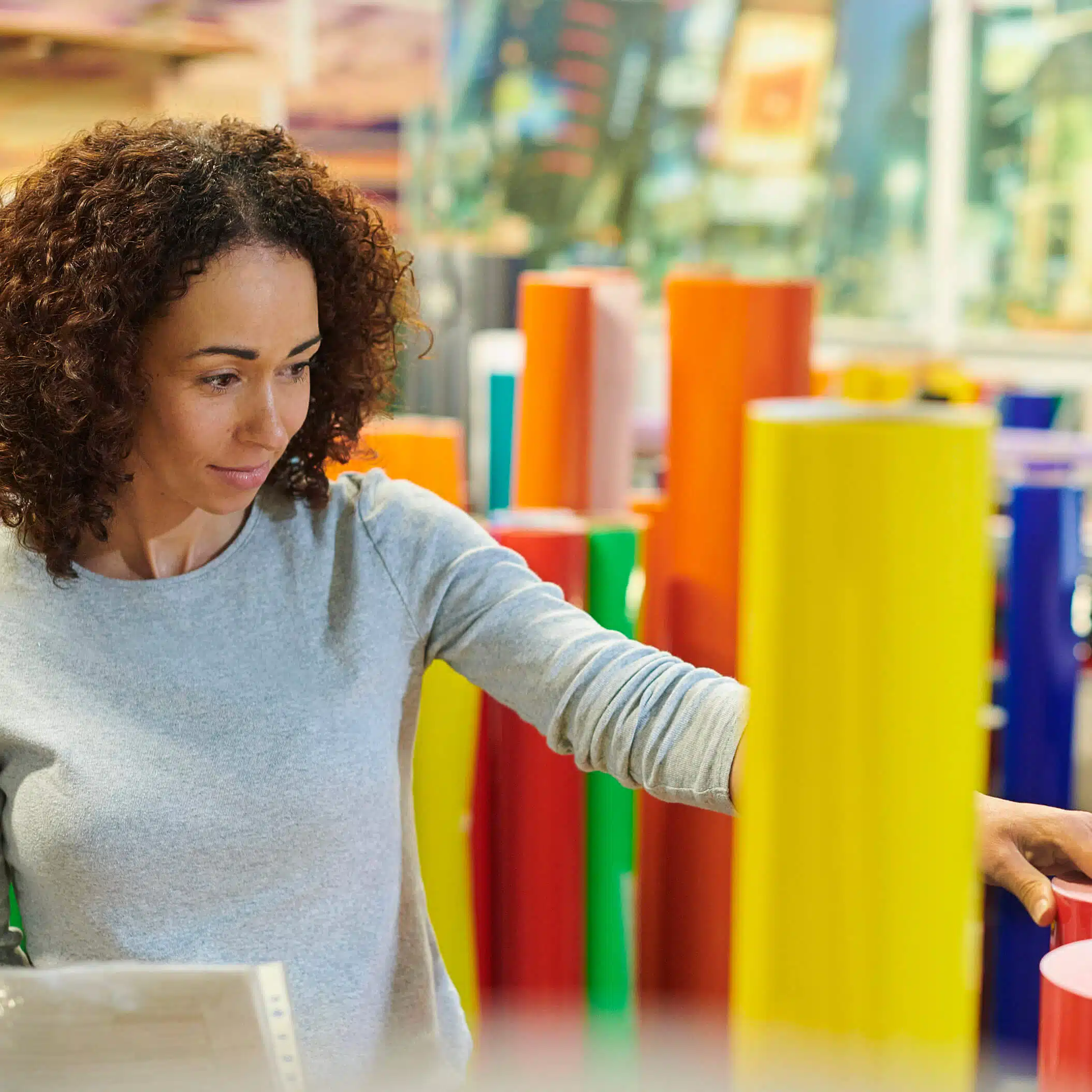 zensmart is showing A woman with curly hair, in a gray long-sleeve shirt, is selecting a colorful roll of material from an array of vibrant rolls meant for signage. She is in a brightly lit store with various colorful rolls around her. The background is slightly blurred. with print workflow automation
