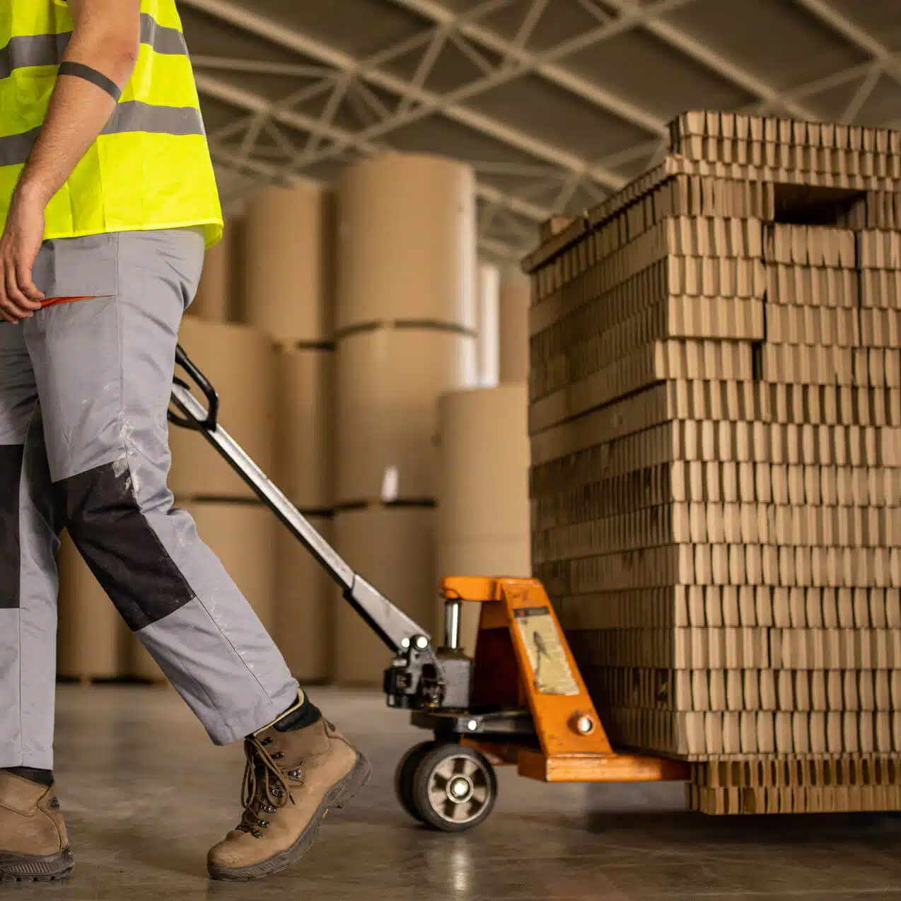 zensmart is showing A worker in a high-visibility yellow vest and gray pants uses a manual pallet jack to move a stack of cardboard boxes adorned with labels in a warehouse. Large rolls of material can be seen in the background amidst industrial surroundings. with print workflow automation