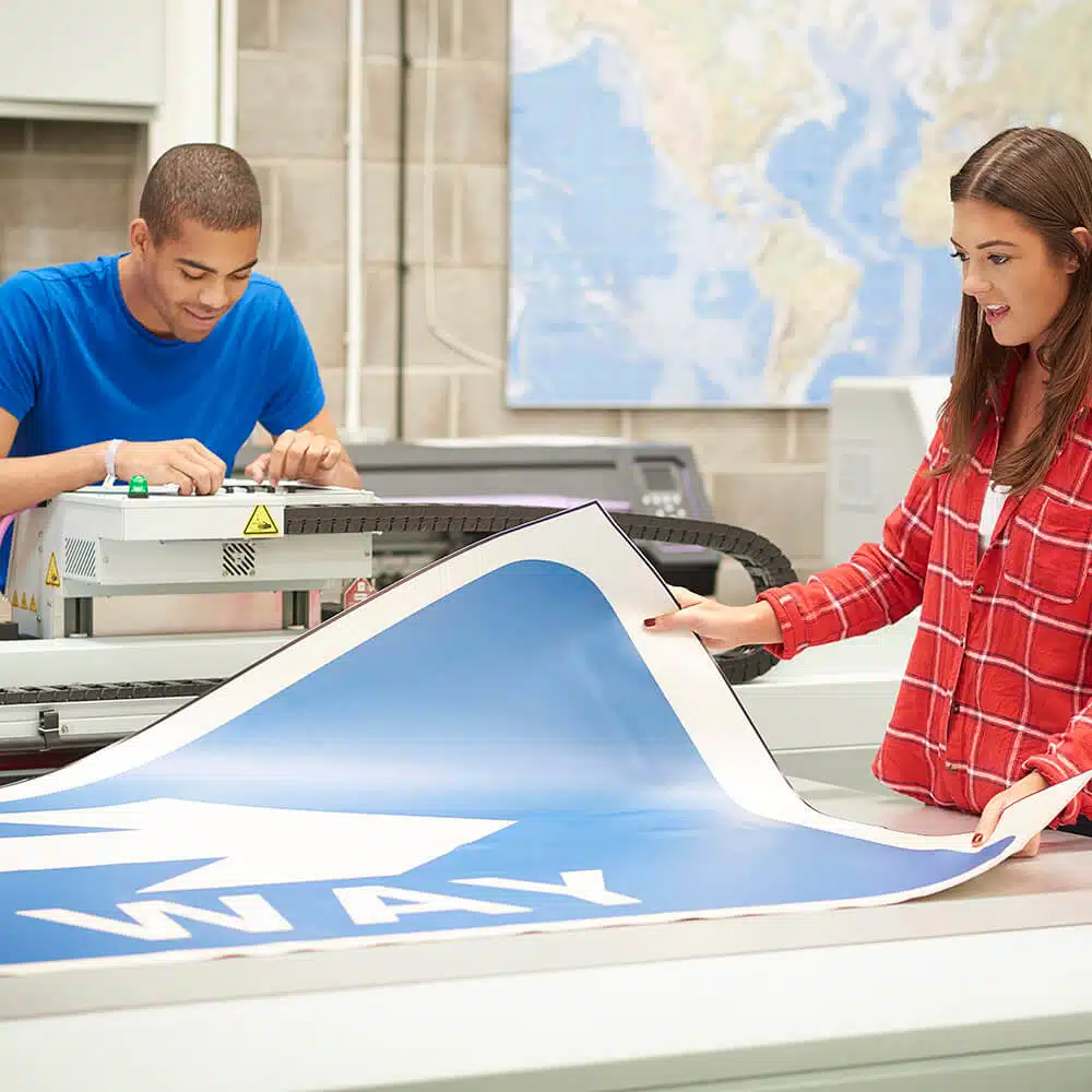 zensmart is showing Two people working in a print shop: one operates a large printer producing a blue banner, while the other, in a red plaid shirt, inspects the printed material. A world map is visible in the background, adding to the global display of their signage expertise. with print workflow automation