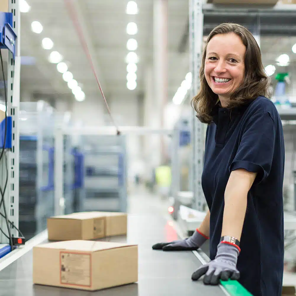 zensmart is showing A woman wearing a dark polo shirt and gloves smiles while standing next to a conveyor belt in a warehouse. Digital print cardboard boxes move along the belt, with industrial shelving and equipment in the background. with print workflow automation