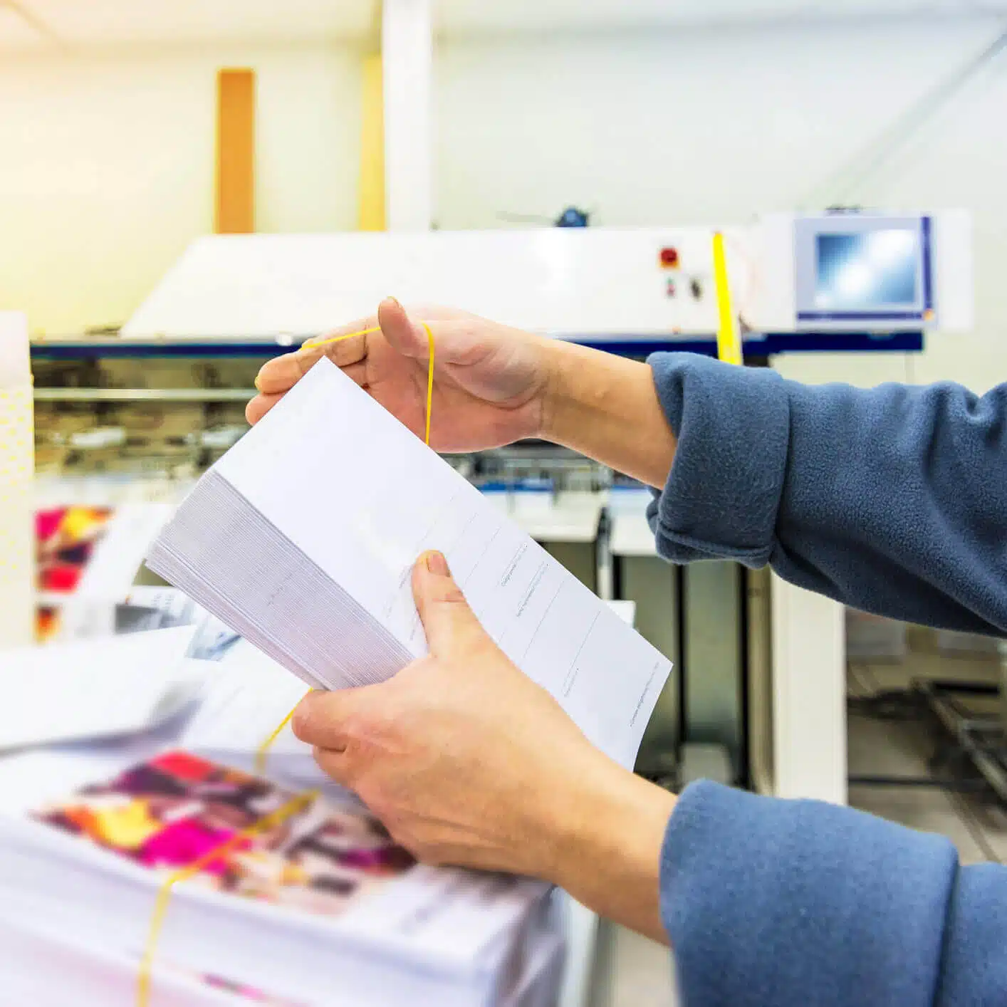 zensmart is showing A person in a blue sleeve is holding a stack of printed papers bound together by a yellow rubber band in a printing facility. The background shows blurred machinery and more stacks of direct mail materials. with print workflow automation